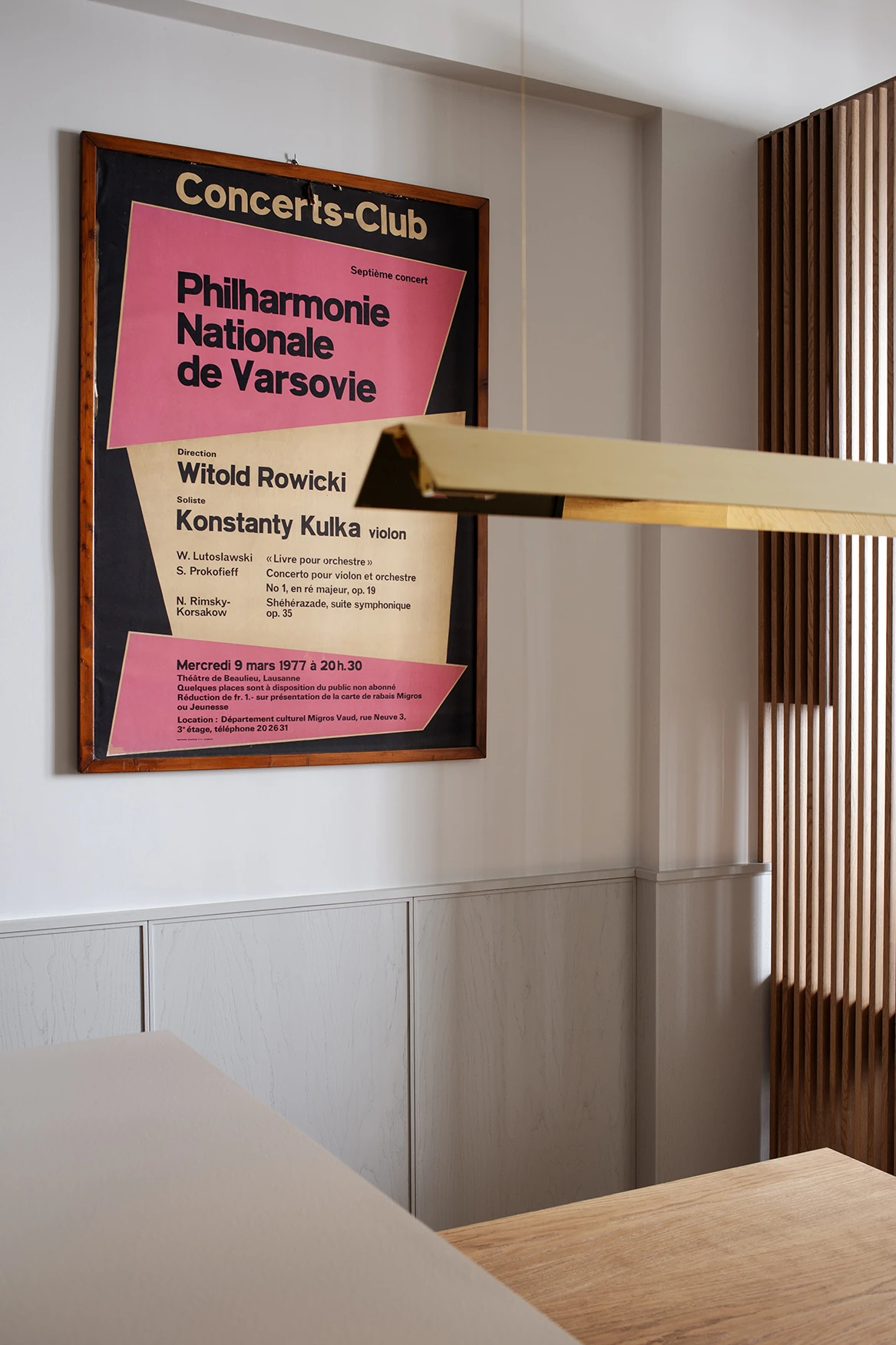 Ściana z fornirowaną lamperią w ciepłym beżu i bezuchwytowymi szafkami, z pionowym ażurem z dębowych listew oraz podłużną mosiężną lampą liniową zawieszoną nad blatem z litego dębu.