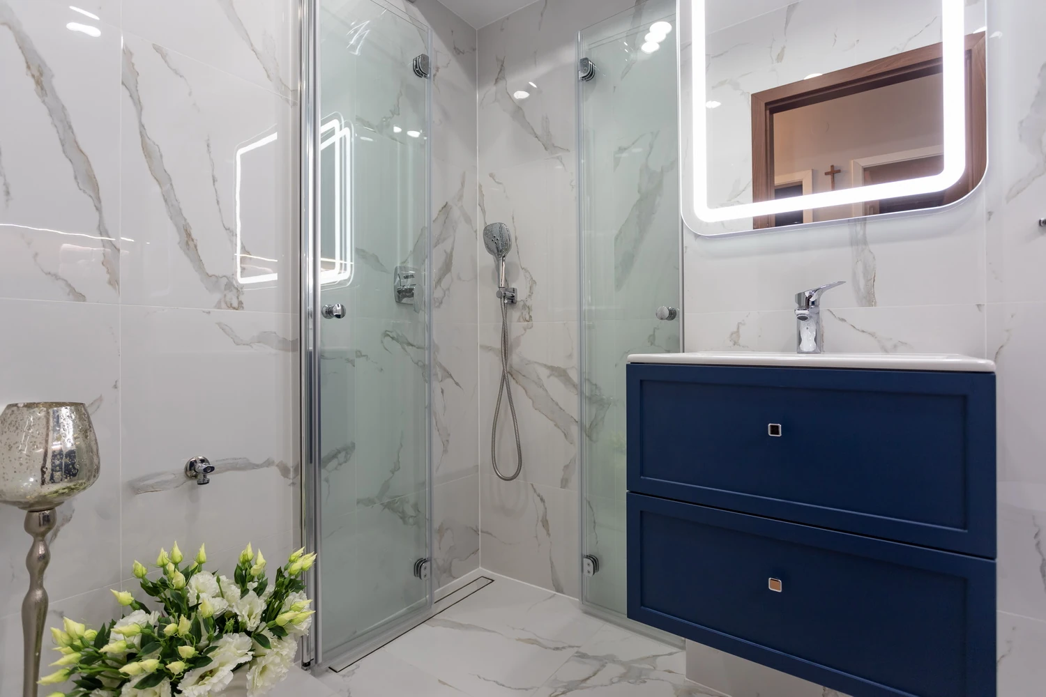 Narożny prysznic walk-in ze szklanymi drzwiami na tle marmuru Calacatta, obok granatowa szafka podumywalkowa z podświetlanym lustrem LED.